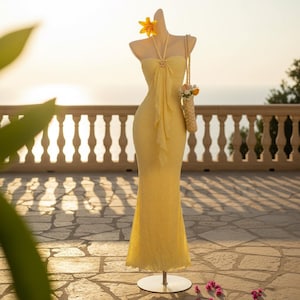 May include: A yellow, floor-length dress on a mannequin. The dress has a halter neckline and a flowing, ruffled design down the front. A small straw bag with flowers hangs from the mannequin's shoulder. A yellow flower adorns the mannequin's head.