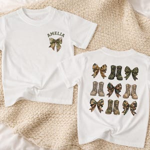 May include: Two white short-sleeved t-shirts. The first shirt features a small camouflage bow graphic above the name AMELIA. The second shirt displays a large grid pattern of camouflage bows and pairs of camouflage boots printed on the back.