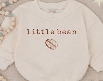 May include: A cream-colored long-sleeve sweatshirt romper featuring the text "little bean" in brown lettering above a brown line-art illustration of a single coffee bean. The garment has a crew neckline and a rounded, bubble-style silhouette.