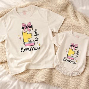 May include: A cream-colored short-sleeve t-shirt and a matching short-sleeve bodysuit, both featuring a graphic of the letter E designed as a yellow pencil with a pink bow, the text "E is for" and the name "Emma" in black script lettering.