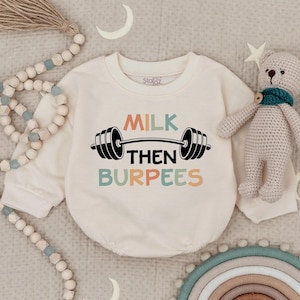 May include: Cream-colored baby onesie with the text "MILK THEN BURPEES" in colorful letters and a barbell graphic. A small teddy bear and decorative beads are also in the image.