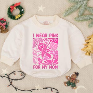 May include: Cream-colored baby bodysuit with long sleeves and a round neckline. The front features a pink floral design and the text "I WEAR PINK FOR MY MOM". Festive decorations surround the bodysuit.