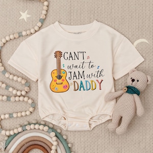May include: Cream-colored baby onesie with a guitar graphic and the text "Can't wait to JAM with DADDY" in colorful letters. The onesie has short sleeves and a round neckline. A small teddy bear is next to the onesie.