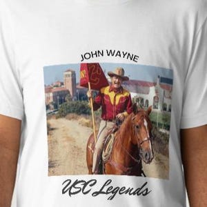 May include: White t-shirt with a graphic of John Wayne on horseback, holding a flag. The image includes the text "JOHN WAYNE" above and "USC Legends" below. The image on the shirt is a vintage photograph.