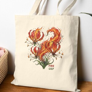 Könnte beinhalten: Eine cremefarbene Tragetasche mit einer lebendigen Illustration einer Feuerlilie. Das Kunstwerk zeigt feurige rote und orangefarbene Blüten mit grünem Laub, und die Worte "FIRE LILY" sind klein gedruckt.