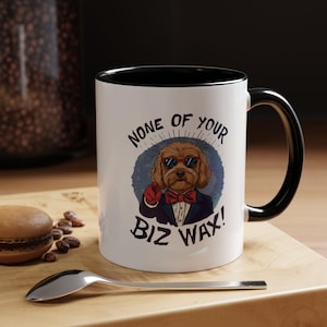 Op de afbeelding: Een witte keramische mok met een zwarte handgreep en binnenrand. De voorzijde toont een afbeelding van een bruine hond met een zonnebril, een rode vlinderdas en een donker pak, die naar voren wijst. De tekst luidt NONE OF YOUR BIZ WAX in zwarte letters.