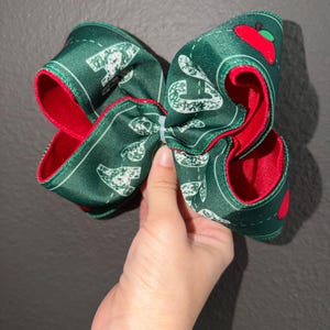 May include: A large hair bow made of dark green ribbon with a red interior lining. The ribbon features a repeating white pattern of school-related icons, including apples and pencils, and is secured in the centre with a small white plastic zip tie.