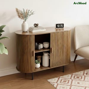 May include: A wooden cabinet with a light brown finish and vertical ribbed detailing. The cabinet has two doors and a shelf inside, displaying various decorative items. A digital clock displays the time 10:35. The cabinet is part of a living room setting.