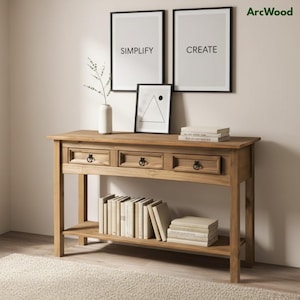 May include: A light brown wooden console table with three drawers and a lower shelf filled with books. Above the table are framed prints with the words "SIMPLIFY" and "CREATE". A white vase and framed art piece are on the table.