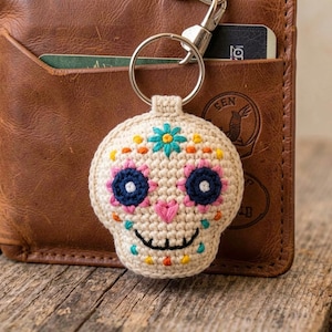 May include: A crocheted skull-shaped keychain with a metal ring. The skull is off-white with colorful embroidered details, including blue, pink, orange, and yellow flowers. The keychain is attached to a brown leather wallet.