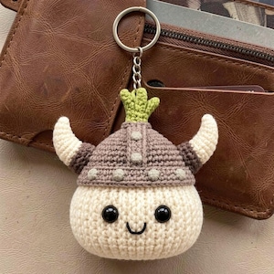 May include: A handmade crochet keychain featuring a smiling, round character wearing a Viking helmet. The helmet is brown and gray, with white and brown horns. The keychain has a metal ring and chain.