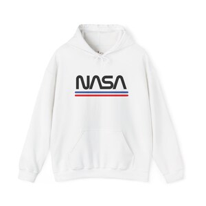Puede incluir: Sudadera con capucha blanca con el logo de la NASA en negro, con rayas rojas y azules debajo. La sudadera tiene un bolsillo delantero y una capucha con cordón. Prenda casual y cómoda.