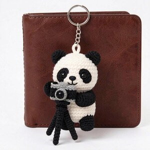 May include: A handmade crochet panda keychain with a silver keyring. The panda is black and white, holding a gray camera on a black tripod. The background is a brown leather wallet.