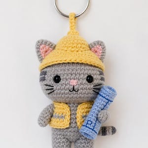 May include: A crocheted cat keychain with a yellow hat and vest, holding a blue scroll. The gray cat has black eyes, whiskers, and a pink nose. The keychain has a silver ring.