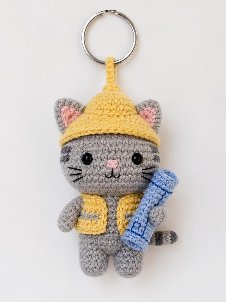 May include: A crocheted cat keychain with a yellow hat and vest, holding a blue scroll. The gray cat has black eyes, whiskers, and a pink nose. The keychain has a silver ring.