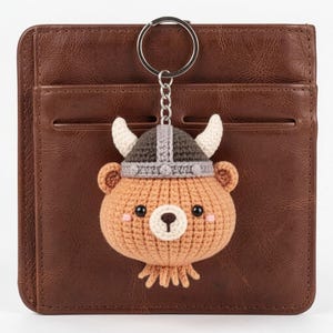 May include: A crocheted bear keychain with a Viking helmet, featuring a brown body, tan snout, and pink cheeks. The keychain is attached to a silver ring and hangs against a brown leather wallet.