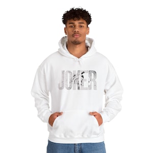 May include: White hooded sweatshirt with the word "JOKER" printed across the chest in a distressed, gray font. The design features a graphic element within the letters. The sweatshirt has a front pocket and a drawstring hood.