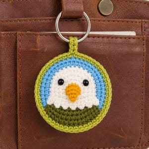 May include: A round crochet keychain featuring a stylized bald eagle face with a white head, a yellow beak, black bead eyes, and a dark olive green chest, all set against a light blue background and bordered by a bright lime green crochet edge.