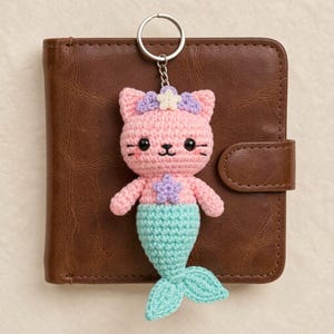 Può includere: Un portachiavi all'uncinetto a forma di testa di gatto rosa con occhi neri, baffi e una piccola bocca. Il gatto indossa un accessorio a stella viola e bianco sulla testa e una stella viola sul petto. Presenta una coda da sirena verde menta.