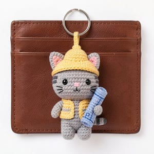 May include: A crocheted cat keychain with a yellow hat and vest, holding a blue scroll. The gray cat has pink inner ears and black whiskers. The keychain is attached to a silver ring and set against a brown leather wallet.