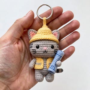 May include: Crocheted gray cat keychain wearing a yellow construction hat and vest, holding a blue blueprint. The cat has black whiskers, pink inner ears, and a striped tail. A silver key ring is attached to the hat.