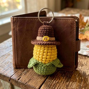 May include: A handmade crochet keychain shaped like a corn cob wearing a pilgrim hat. The keychain is yellow, brown, green, and tan. The keychain is attached to a silver metal ring. The keychain is next to a brown leather wallet.