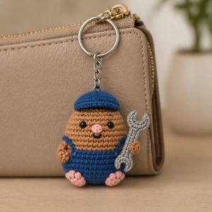 Puede incluir: Un llavero de ganchillo hecho a mano con un personaje de mecánico marrón, azul y rosa que sostiene una llave inglesa gris. El personaje lleva una gorra azul y un mono. El llavero está unido a un anillo plateado y a una cartera de cuero beige.