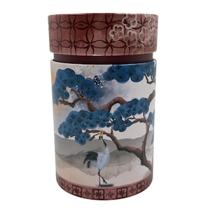 Puede incluir: Un recipiente cilíndrico con tapa. El recipiente presenta una ilustración detallada de una grúa, pinos azules y un paisaje montañoso. La tapa tiene un estampado floral y un diseño geométrico en tonos marrón y rosa.