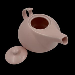 Könnte beinhalten: Eine hellrosa Keramik-Teekanne mit passendem Deckel. Die Teekanne hat einen runden Körper, einen gebogenen Griff und einen Ausguss. Der Deckel hat einen kleinen Knopf und ein Loch. Der Hintergrund ist schwarz.
