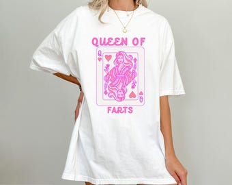 Può includere: Una t-shirt bianca a maniche corte con una grafica rosa di una carta da gioco raffigurante una regina. Il testo QUEEN OF FARTS è stampato in rosa sopra e sotto il disegno della carta. La maglietta ha una vestibilità ampia e girocollo.