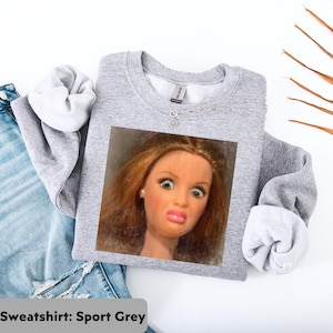 May include: A light gray sweatshirt with a printed image of a doll's face making a surprised expression. The sweatshirt is folded, with the sleeves showing a white lining. The text "Sweatshirt: Sport Grey" is at the bottom.