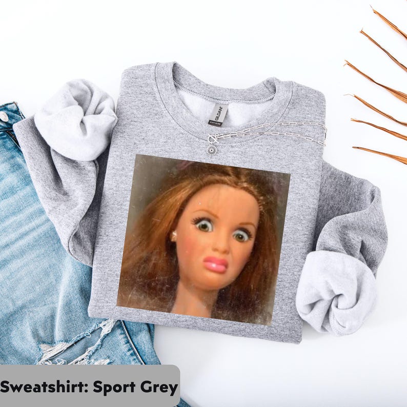 May include: A light gray sweatshirt with a printed image of a doll's face making a surprised expression. The sweatshirt is folded, with the sleeves showing a white lining. The text "Sweatshirt: Sport Grey" is at the bottom.