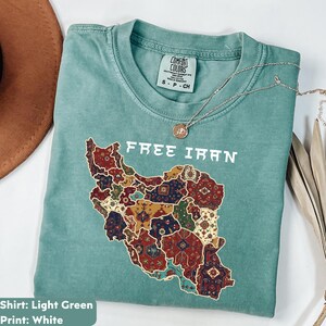 以下が含まれることがあります： イランの地図の上に「FREE IRAN」の文字がプリントされた、明るい緑色のTシャツ。地図はカラフルなカーペットのデザインで埋められています。シルバーのネックレスとペンダントがシャツの上にあります。「Comfort Colors」とラベルされており、サイズはSです。