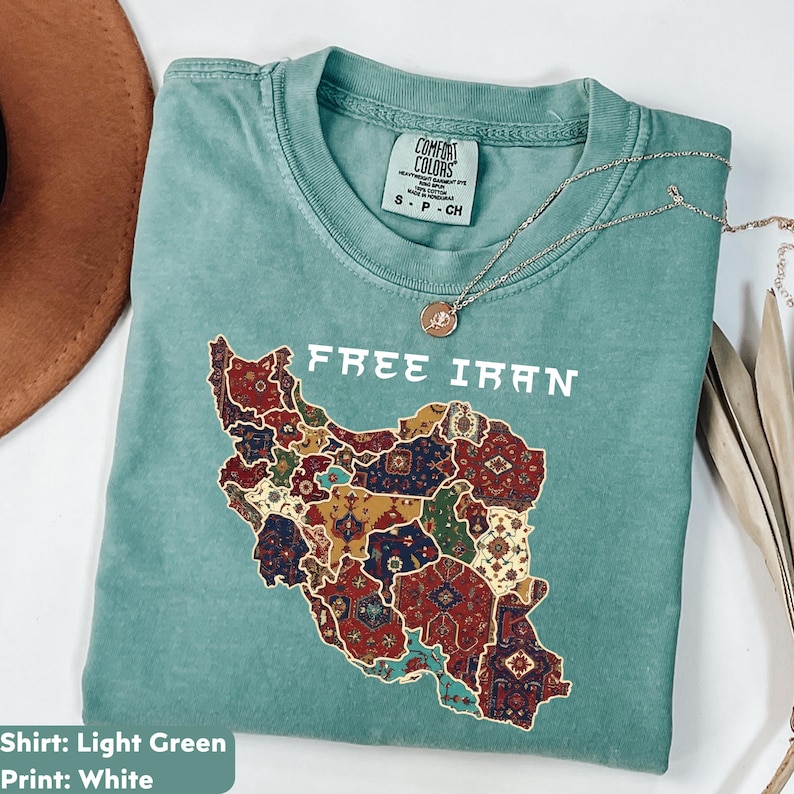 以下が含まれることがあります： イランの地図の上に「FREE IRAN」の文字がプリントされた、明るい緑色のTシャツ。地図はカラフルなカーペットのデザインで埋められています。シルバーのネックレスとペンダントがシャツの上にあります。「Comfort Colors」とラベルされており、サイズはSです。