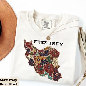 以下が含まれることがあります： 「FREE IRAN」の文字と、カラフルな模様で装飾されたイランの地図がプリントされたアイボリー色のTシャツ。金のネックレスと茶色の帽子も写っています。