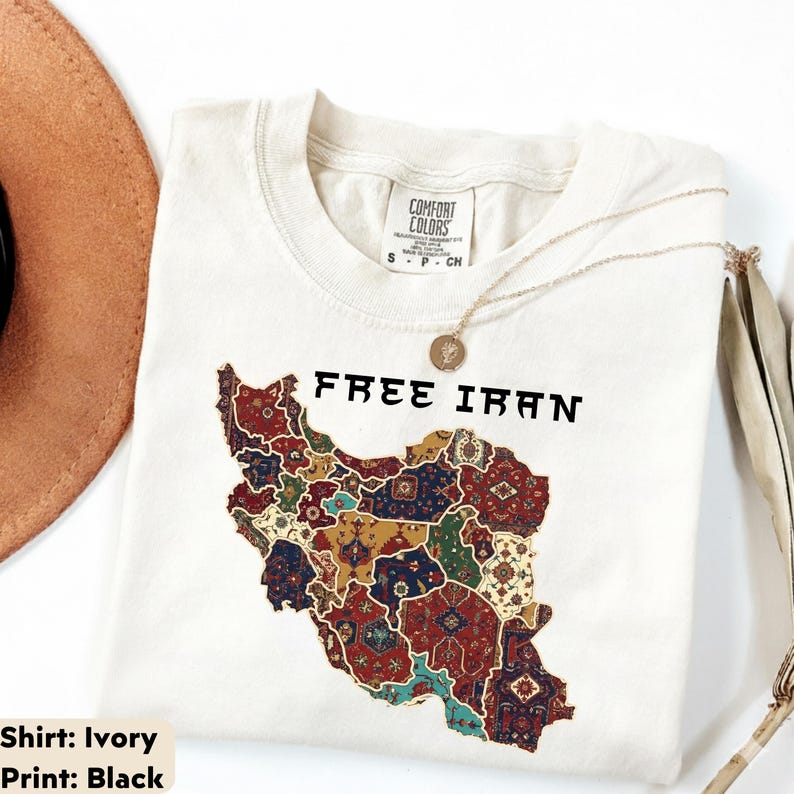 以下が含まれることがあります： 「FREE IRAN」の文字と、カラフルな模様で装飾されたイランの地図がプリントされたアイボリー色のTシャツ。金のネックレスと茶色の帽子も写っています。