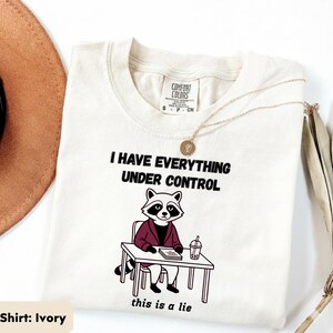 May include: An ivory t-shirt featuring the phrases "I HAVE EVERYTHING UNDER CONTROL" and "this is a lie." A cartoon raccoon in a blazer is depicted at a desk. A gold necklace and a brown hat are also visible.
