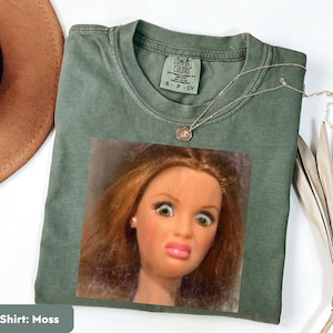 May include: Moss green t-shirt with a doll face graphic. The shirt has a crew neck and short sleeves. A silver necklace with a small pendant is draped across the shirt. The shirt is made of a soft material.