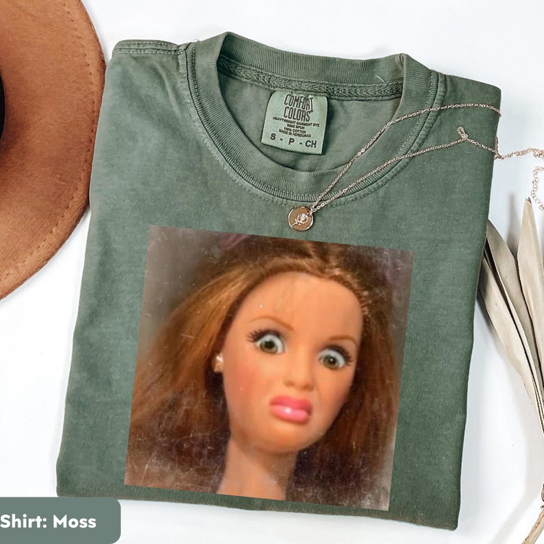 May include: Moss green t-shirt with a doll face graphic. The shirt has a crew neck and short sleeves. A silver necklace with a small pendant is draped across the shirt. The shirt is made of a soft material.