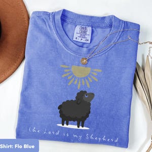 Könnte beinhalten: Flo Blue T-Shirt mit einem schwarzen Schaf und einer goldenen Sonne. Darunter steht der Text "the Lord is my Shepherd". Eine goldene Halskette liegt auf dem gefalteten T-Shirt.