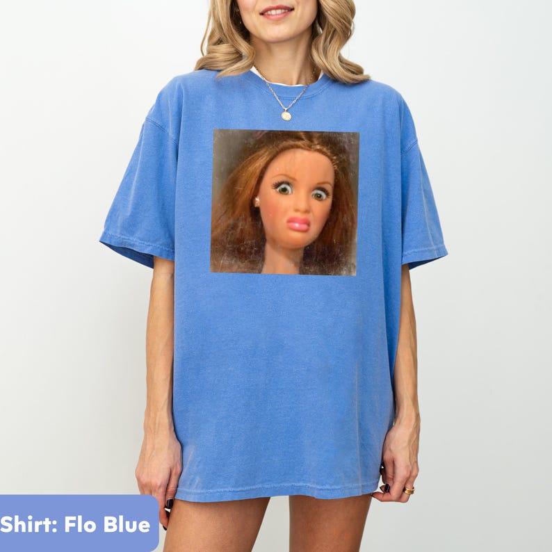 May include: A light blue t-shirt with a graphic of a doll's face making a surprised expression. The doll has brown hair, large eyes, and a puckered mouth. The shirt is labeled "Shirt: Flo Blue".