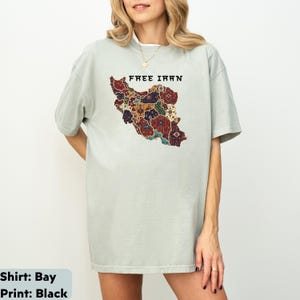 以下が含まれることがあります： 「FREE IRAN」の文字と、イランのカラフルな地図がプリントされた、薄い緑色のTシャツ。 地図のデザインには、赤、青、金など、さまざまな模様と色が使われています。 ゆったりとしたフィット感のシャツです。