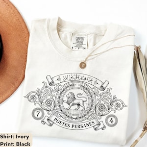 May include: Ivory-colored t-shirt with a black graphic design. The design features a lion inside a wreath, with text in a banner above and below. The shirt also has the numbers 7 and 4 on either side of the design.