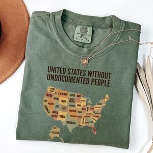 May include: Olive green t-shirt with the text "UNITED STATES WITHOUT UNDOCUMENTED PEOPLE" and a map of the United States with tribal names. A gold necklace with a pendant is draped across the shirt.