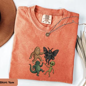 Può includere: Una t-shirt a maniche corte color corallo con etichetta Comfort Colors sul collo. La grafica mostra un Bigfoot marrone, una creatura falena nera, un mostro simile a una lucertola verde e un alieno verde che ballano insieme.