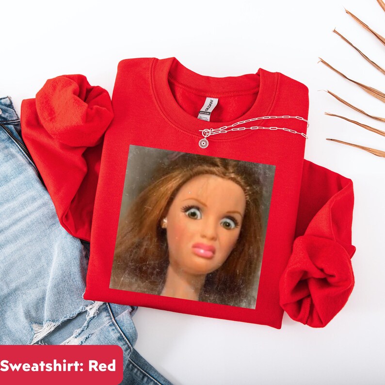 May include: A bright red sweatshirt featuring a framed image of a doll with a surprised expression. A silver necklace with a small pendant is draped on the sweatshirt. The sweatshirt is folded next to a pair of blue jeans.