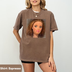May include: Espresso-colored t-shirt featuring a square image of a doll with a surprised expression. The shirt has short sleeves and a crew neck. The model is wearing a white undershirt and black shorts.