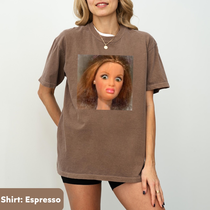 May include: Espresso-colored t-shirt featuring a square image of a doll with a surprised expression. The shirt has short sleeves and a crew neck. The model is wearing a white undershirt and black shorts.