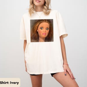 May include: Ivory-colored t-shirt featuring a square image of a doll's face with a surprised expression. The doll has red hair, large eyes, and a pink pout. The shirt is a casual style.