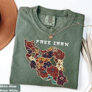 以下が含まれることがあります： モスグリーンのTシャツ。前面には「FREE IRAN」の文字と、カラフルな模様で埋められたイランの地図がプリントされています。シルバーのネックレスがTシャツの上に置かれています。Tシャツには「Comfort Colors」と記載されています。
